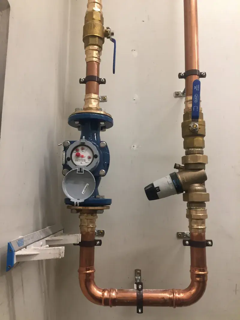 A brass and copper water meter with a testable backflow prevention assembly mounted on a wall
