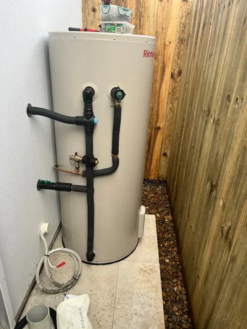 A newly installed Rinnai electric hot water storage tank with black pipework against a timber fence