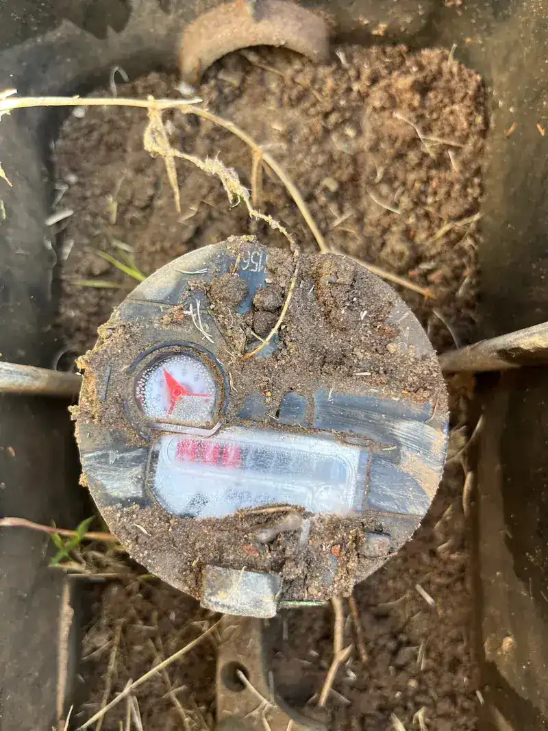 A residential water meter uncovered in the ground, dial visible through the dirt